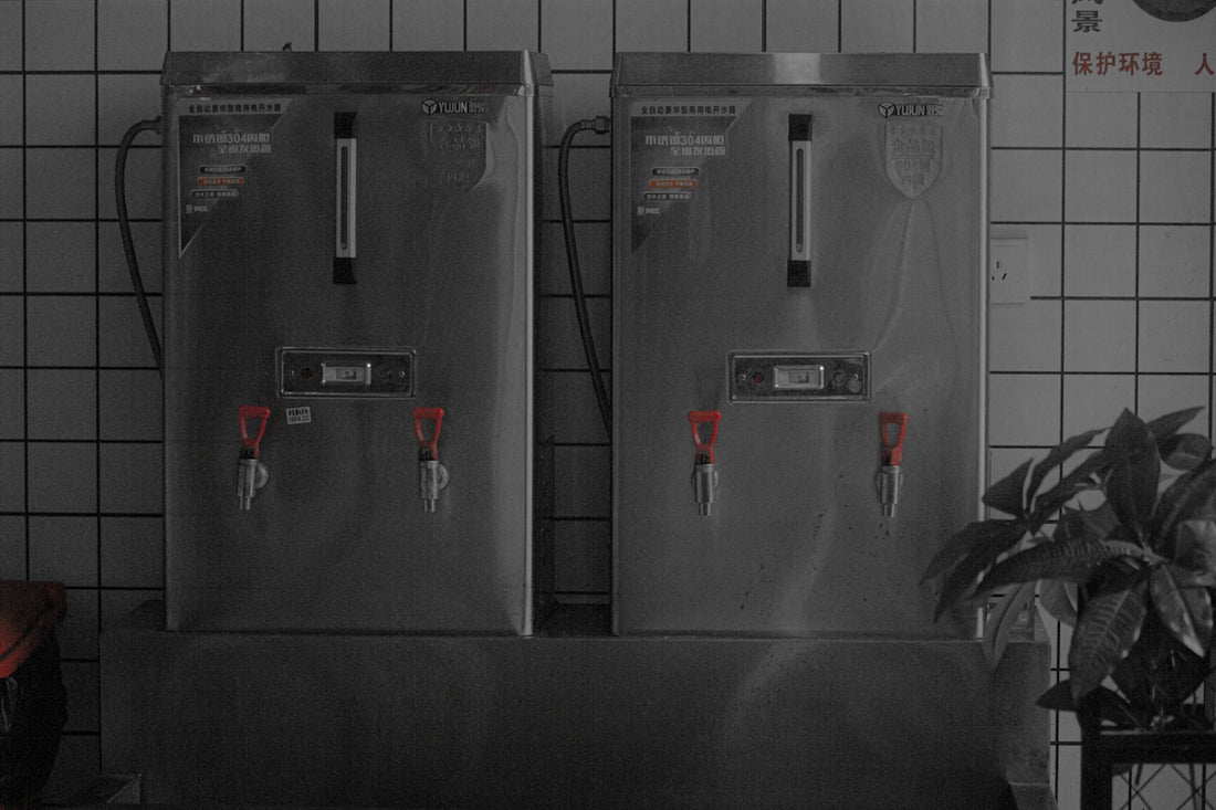 Two stainless steel hot water dispensers on a tiled wall.