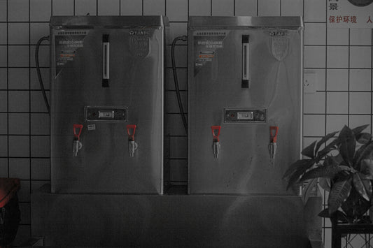 Two stainless steel hot water dispensers on a tiled wall.